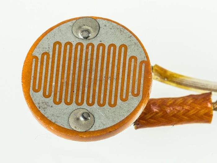 Komponen LDR berbentuk bulat pipih dengan pola jalur resistor zig-zag berwarna oranye di permukaannya.